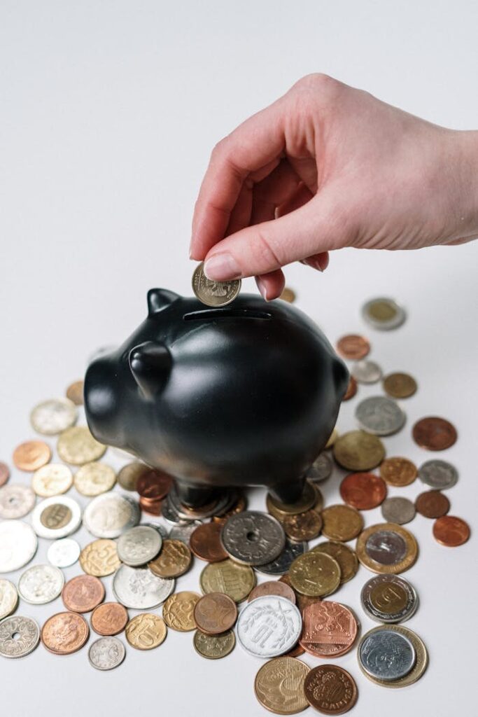 pexels-photo-3943723 Hand inserting coin into a black piggy bank surrounded by various coins, symbolizing savings.