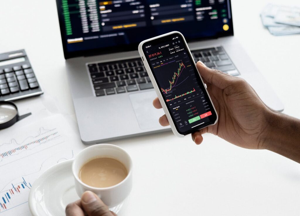 pexels-photo-6801874 A person trades stocks on a smartphone with a laptop, coffee cup, calculator, and documents in view.